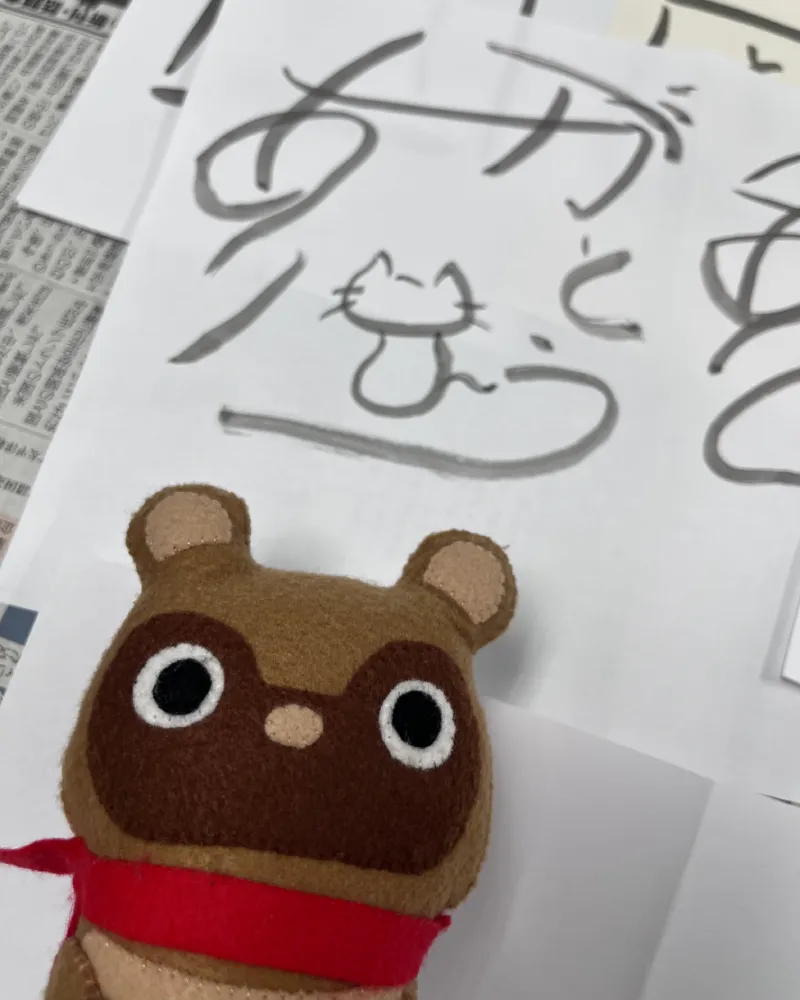 alt="和游字でネコのイラストと『ありがとう』が描かれた作品を前にした、マスコットのたぬきち