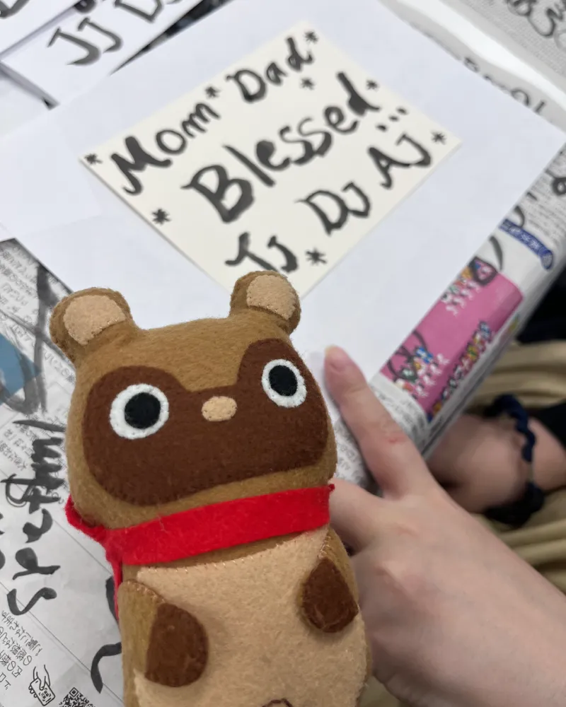 和游字体験で『Mom Dad Blessed Jj. Dj Aj.*』と書かれた筆文字作品を指差す参加者の手と、見守るマスコットキャラクターのたぬきち