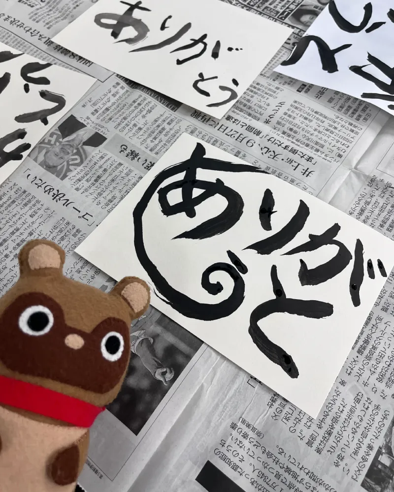 新聞紙の上に並べられた様々なタッチの和游字『ありがとう』と、手前に座るマスコットキャラクターのたぬきち