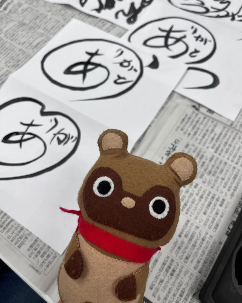 円と組み合わさった和游字『ありがとう』のデザイン練習紙を見つめるマスコットキャラクターのたぬきち