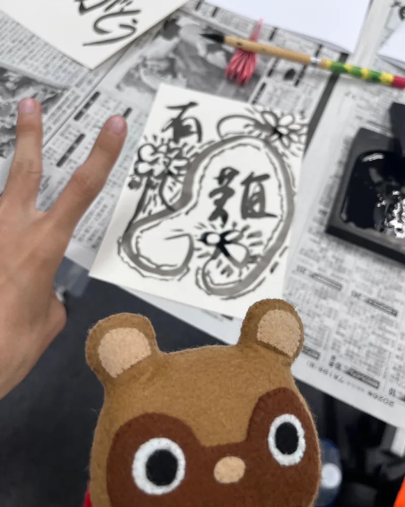 和游字体験にて、漢字の『有難』と花のイラスト、そして英字の『Thank you』がデザインされた芸術的な作品と、ピースサインをする参加者の手、見守るマスコットのたぬきち
