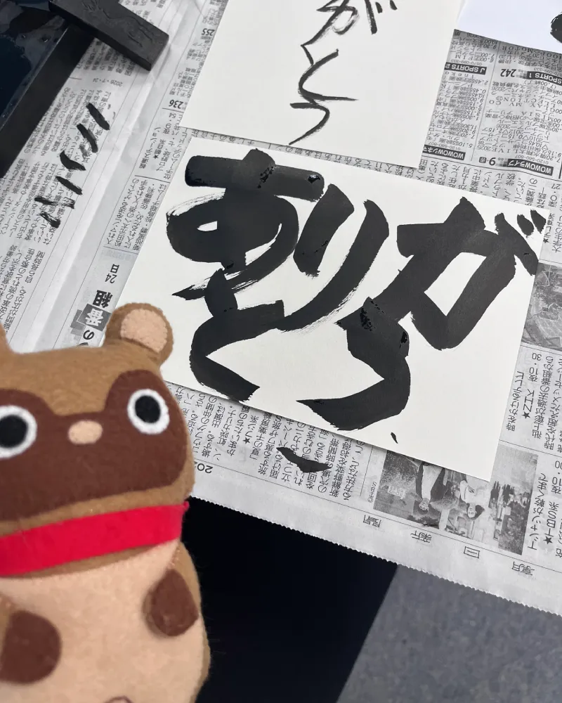 alt="和游字体験にて、太い墨で大胆に書かれた『ありがとう』の作品と、横に置かれた英字『Thank you』の練習紙、そして見守るマスコットキャラクターのたぬきち