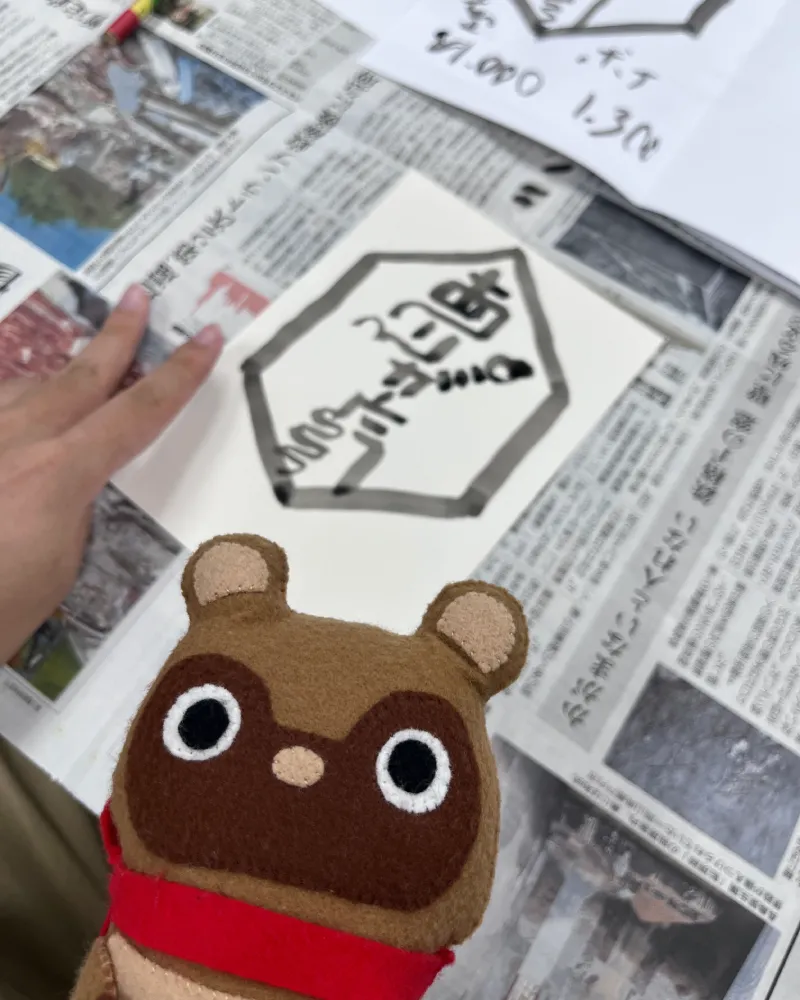和游字体験にて、六角形の枠の中に古代文字のような抽象的な筆文字が描かれたユニークな作品と、それを見つめるマスコットキャラクターのたぬきち